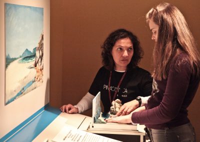 Educadora Kika mediando visita com pessoa com deficiência visual que explora maquete tátil da exposição.