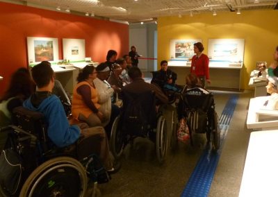 grupo com pessoas em cadeira de rodas esta no centro da exposição recebendo atendimento de Amanda Tojal