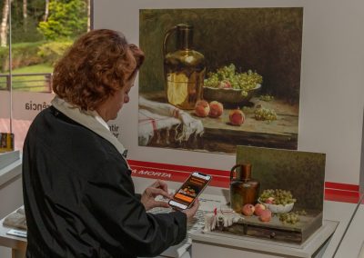 Amanda Tojal segurando celular, na tela o webapp da exposição acessando informações sobre obra uvas e pêssegos de Pedro Alexandrino