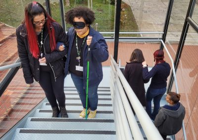 Educadores em dupla, uma guiando e outra simulando pessoa com deficiência visual, olhos vendados e bengala, todos sobrem escadas.
