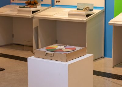 Foto colorida na vertical do jogo roleta das cores sobre uma base quadrada, ao fundo duas bancadas da exposição com tema marinha.