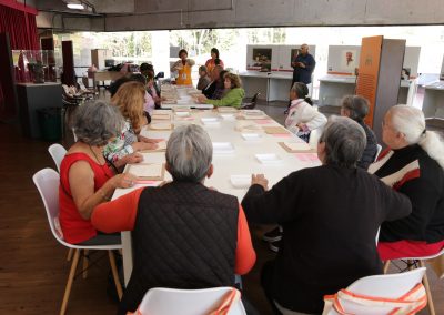 Grupo de idosos sentados ao redor de uma grande mesa com materiais da oficina de sacolas, escutam orientações de Amanda Tojal.