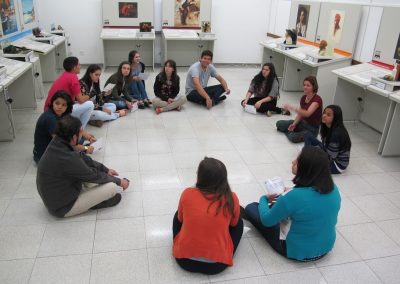 Grupo de estudantes e Amanda, sentados em círculo no chão da exposição.